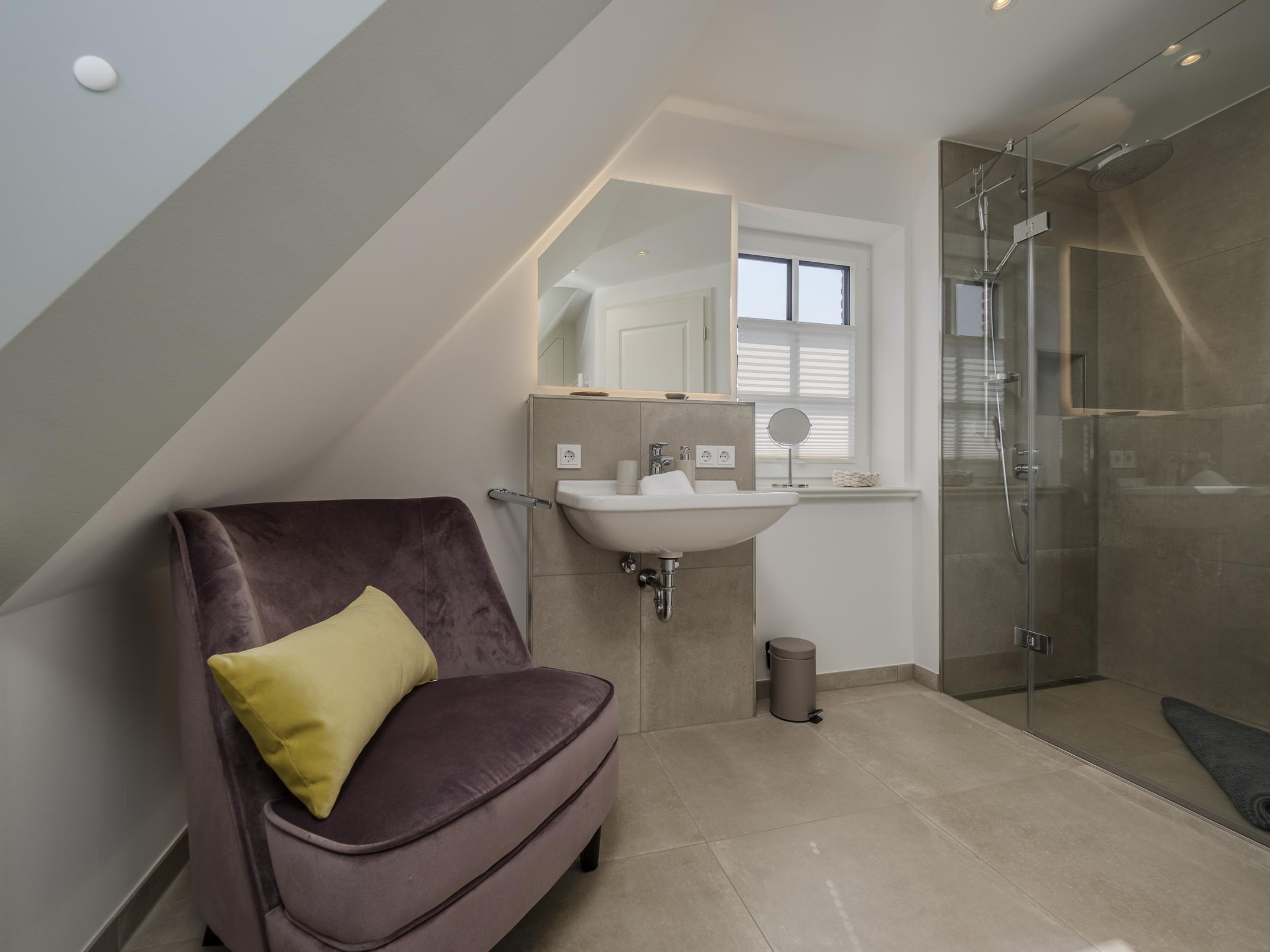 Geräumiges Wellness-Bad mit bodengleicher Regendusche und gemütlichem Samtsessel.