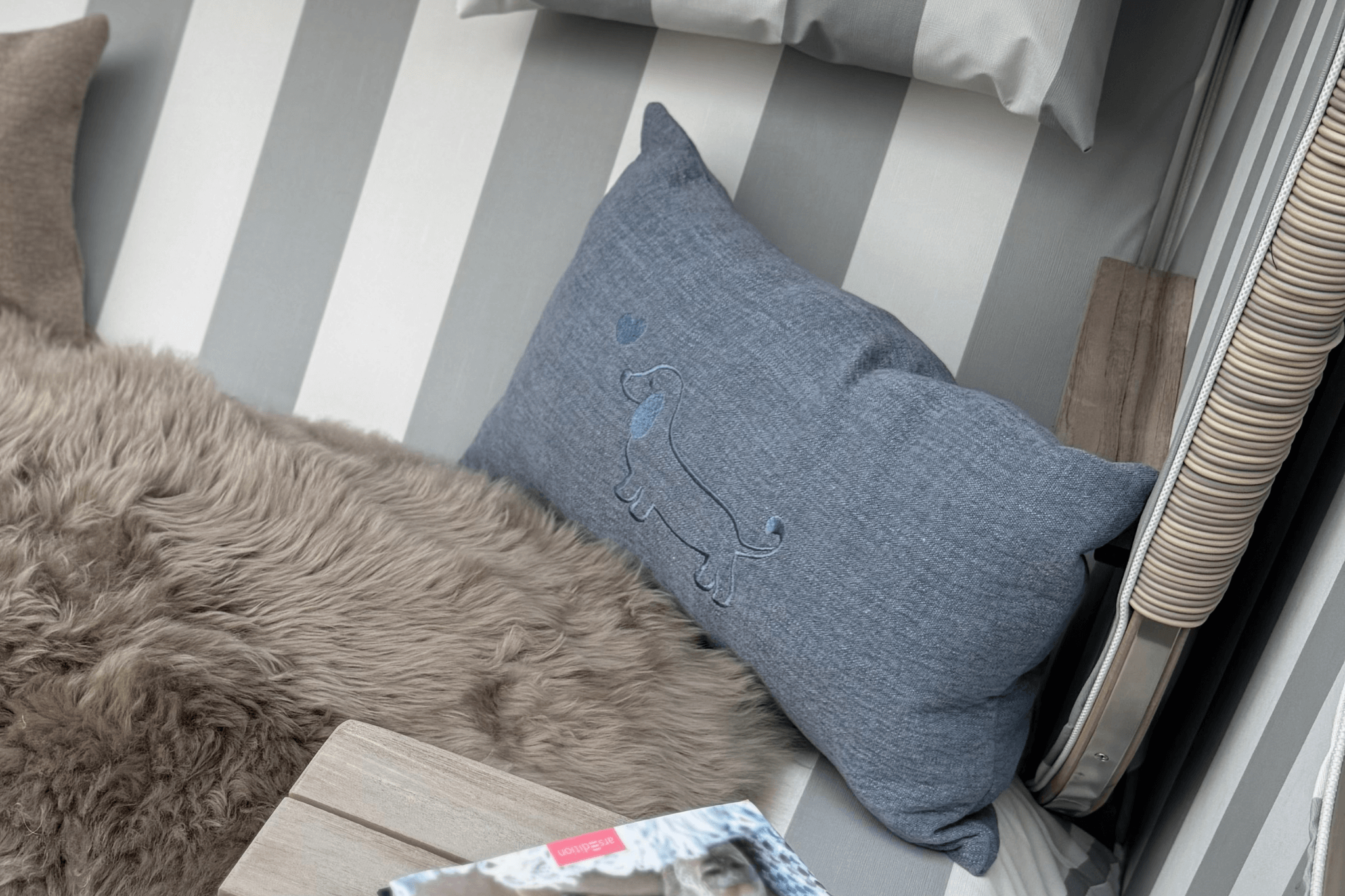 Greta & PAUL Leinen-Kissen in Hellblau mit Dackel-Stickerei, stilvoll auf einem flauschigen Fell in einem Strandkorb arrangiert.