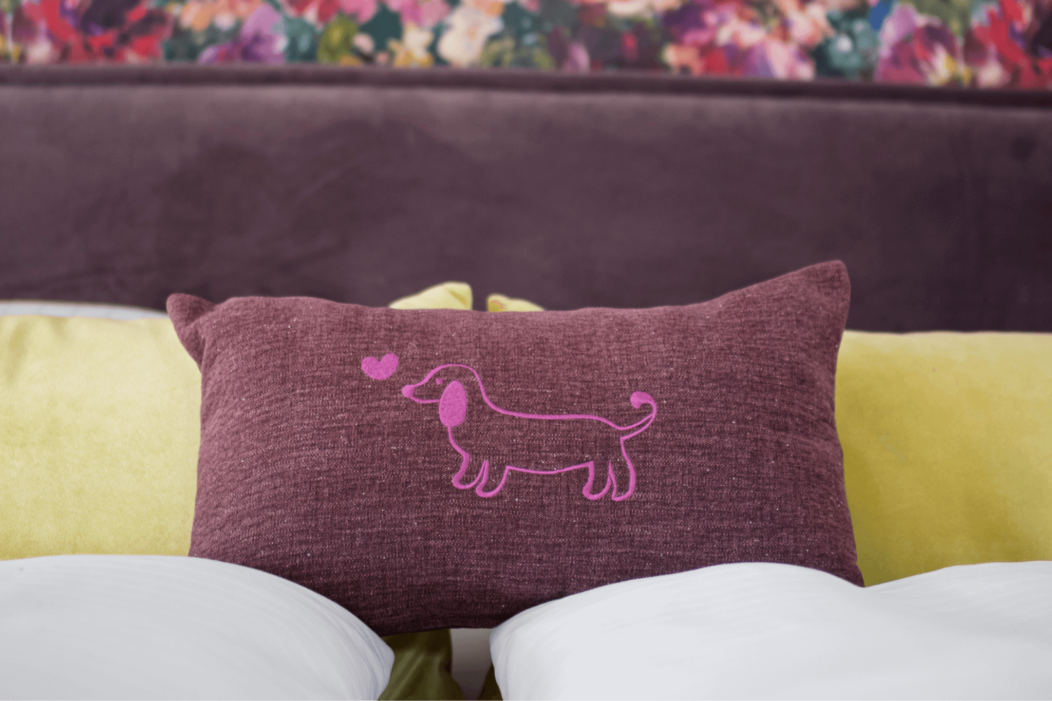 Nahaufnahme des Greta & PAUL Leinen-Kissens in Lila mit leuchtend pinker Dackel-Stickerei, platziert auf einem Bett.