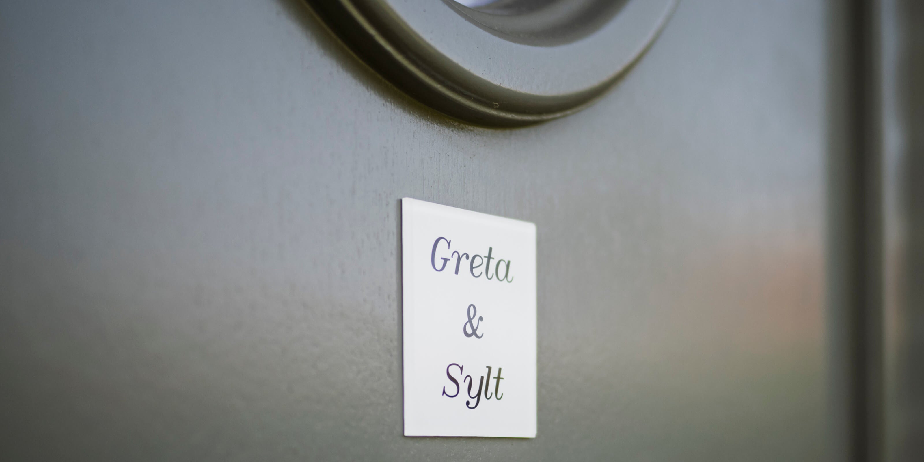 Nahaufnahme des 'Greta & Sylt' Schildes an der Eingangstür des Ferienhauses, das Gäste herzlich willkommen heißt.