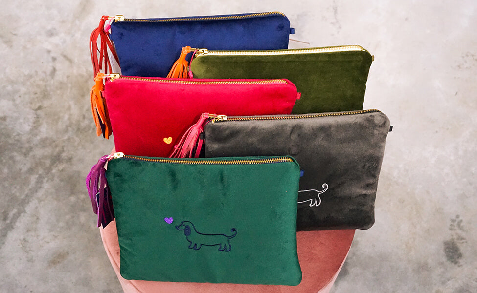 Verschiedene Samt-Clutches Stella von Greta & PAUL, in Dunkelblau, Dunkelgrün, Anthrazit, Pink und Olivgrün mit Dackel- und Herz-Bestickung.
