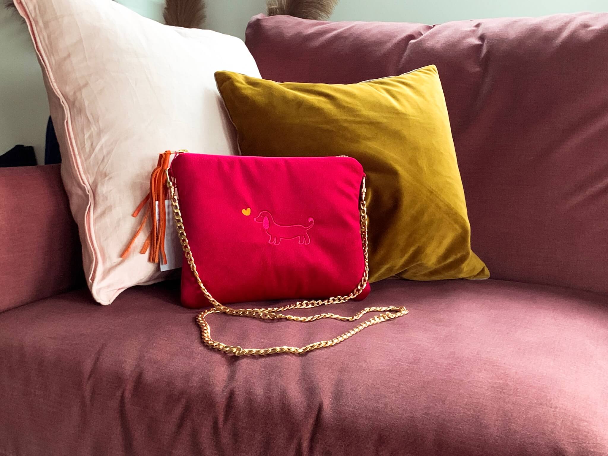 Greta & PAUL Clutch 'Stella' in Pink mit Dackel-Stickerei, stilvoll auf einem Sofa zwischen bunten Kissen arrangiert.
