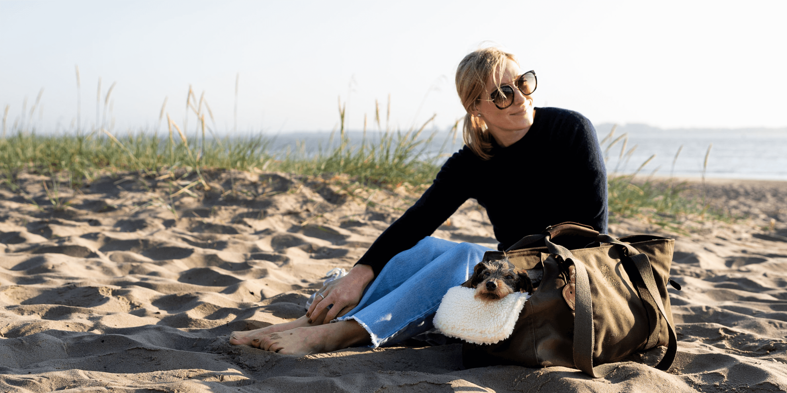 Julia sitzt mit ihrer Dackeldame Gretchen am Stader Strand in einer handgefertigten Hundetasche von Greta & PAUL mit einem weichen Teddyfell-Inlay.