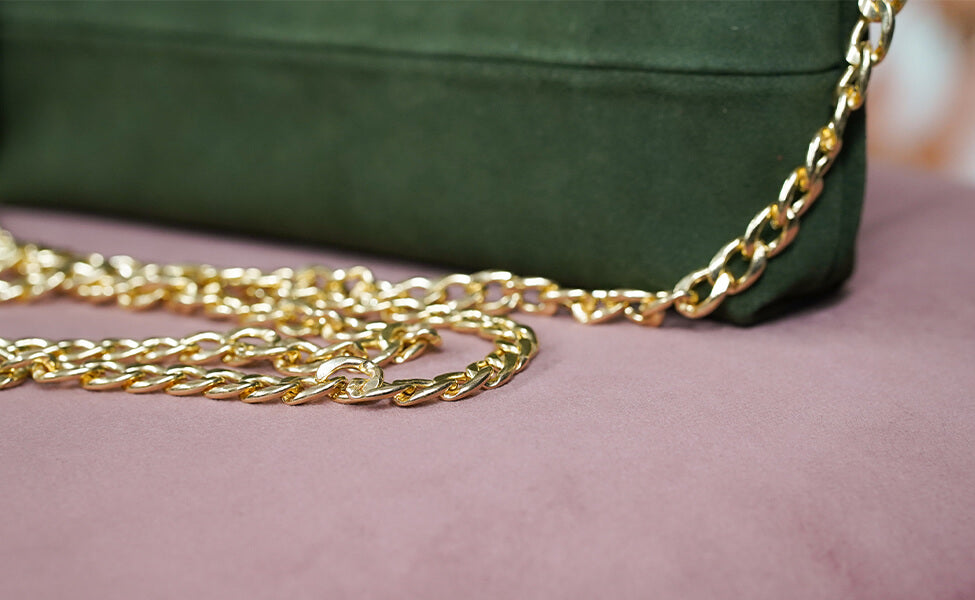 Nahaufnahme der goldenen Schulterriemen-Kette der Greta & PAUL Wildleder-Clutch Stella Deluxe auf rosa Untergrund.